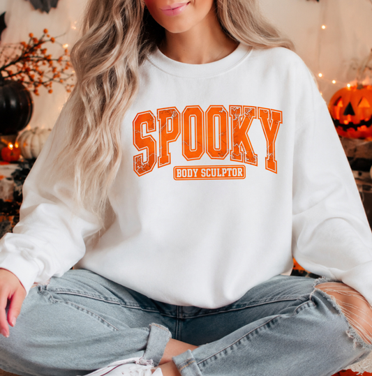 A woman wearing a white crewneck sweatshirt with 'SPOOKY BODY SCULPTOR' printed in orange across the front, paired with ripped light-wash jeans, set against a cozy, warmly lit Halloween-themed background with pumpkins and string lights. Sweatshirt for body contouring specialists.