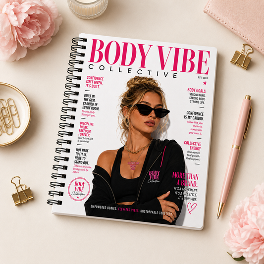 A spiral-bound notebook styled like a magazine cover titled 'BODY VIBE COLLECTIVE' is placed on a light surface with pink flowers, gold paper clips, a pink pen, and a small glass dish around it. The cover shows a woman in sunglasses and a black top with pink text and graphics. Notebooks, clothing and accessories for beauty professionals.