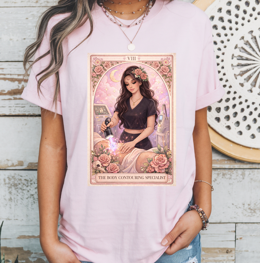 A woman wearing a light pink t-shirt with a large tarot card graphic on the front. The graphic depicts 'The Body Contouring Specialist' card with a woman in a black dress surrounded by pink roses.