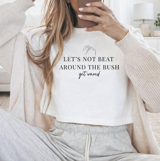A woman wearing a white cropped graphic t-shirt with the text 'LET'S NOT BEAT AROUND THE BUSH get waxed' in black print, styled with a beige cardigan and gray sweatpants in a cozy indoor setting. Novelty shirts and apparel for estheticians, body contouring specialists, and beauty professionals.
