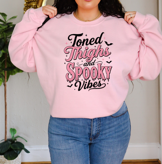 A person wearing a light pink crewneck sweatshirt with the text 'Toned Thighs and Spooky Vibes' in black and pink lettering, paired with blue jeans. Sweatshirt for body contouring specialists.
