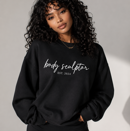 A woman with curly black hair wearing a black crewneck sweatshirt with 'body sculptor EST. 2024' printed in white script across the chest, posed against a neutral gray background. Sweatshirt for body contouring specialists.