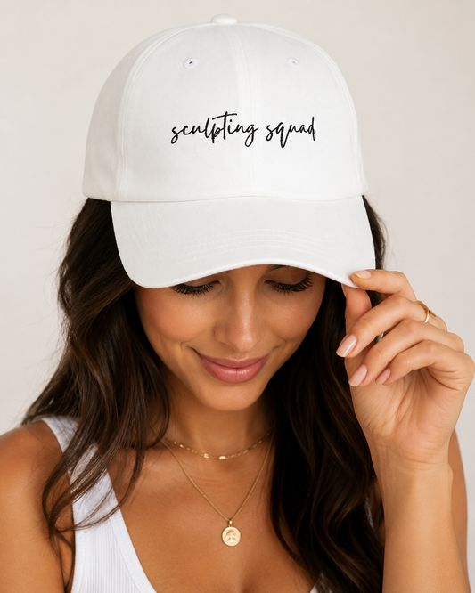 A woman wearing a white baseball cap with 'sculpting squad' embroidered in black script on the front, photographed against a soft light beige background. Hats, accessories and apparel for body contouring specialists.