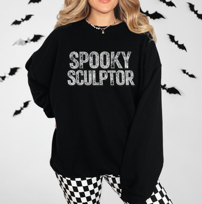 A woman wearing a black oversized sweatshirt with 'SPOOKY SCULPTOR' printed in distressed white letters across the chest, paired with black-and-white checkered pants against a white background with hanging black bats. Halloween sweatshirt for a body sculptor.