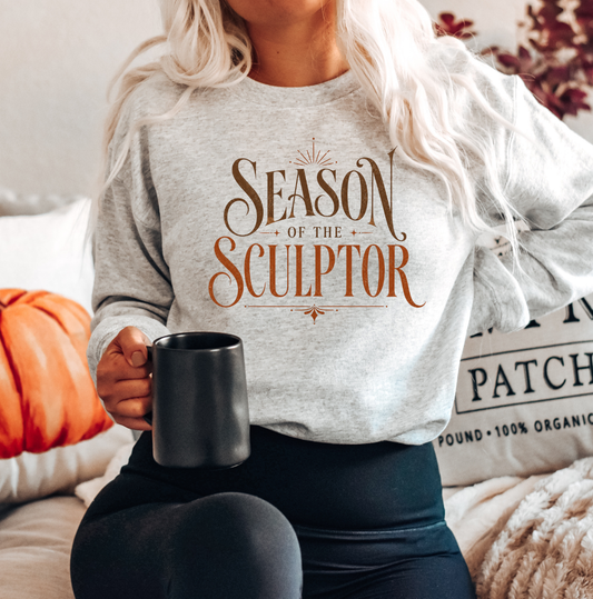 A woman wearing a light heather gray long-sleeve crewneck sweatshirt with the text 'SEASON OF THE SCULPTOR' in brown, holding a black mug while sitting on a couch. Clothing and apparel for body contouring specialists.