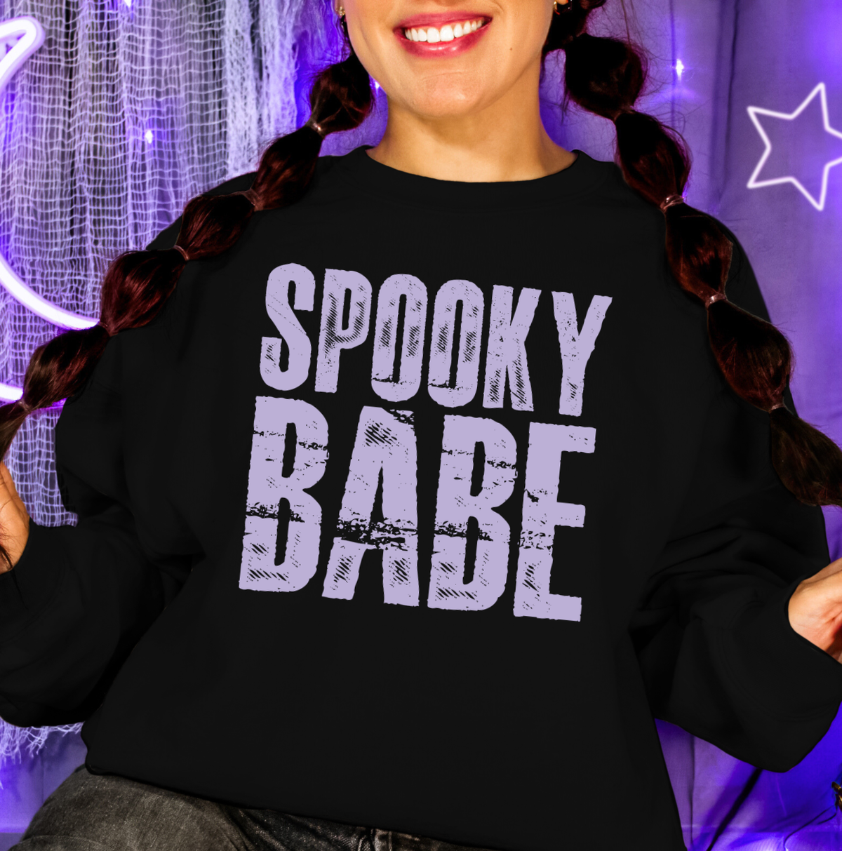 A person wearing a black crewneck sweatshirt with a distressed light purple graphic that reads 'SPOOKY BABE'. The background features purple lighting, a star-shaped neon light, and a net curtain. Halloween graphic shirts and sweatshirts for women.