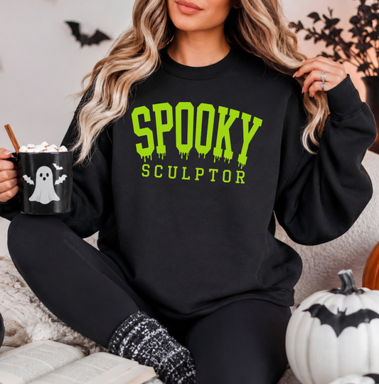 A woman wearing a black crewneck sweatshirt with 'SPOOKY SCULPTOR' printed in bright green dripping letters, sitting in a cozy indoor setting and holding a black mug with a ghost graphic. Sweatshirt for body contouring specialists and body sculptors.