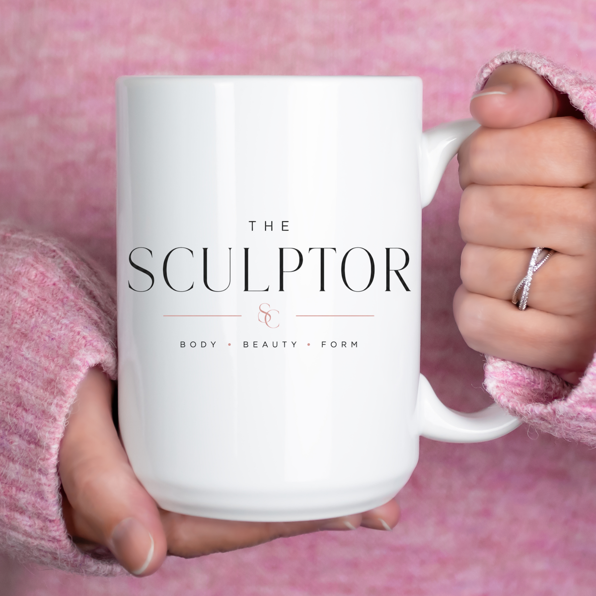 A white ceramic mug with the text 'THE SCULPTOR & BODY • BEAUTY • FORM' printed on the front, held by two hands wearing pink knit sleeves against a pink textured background. Clothing, apparel, and accessories for body contouring specialists and beauty professionals.