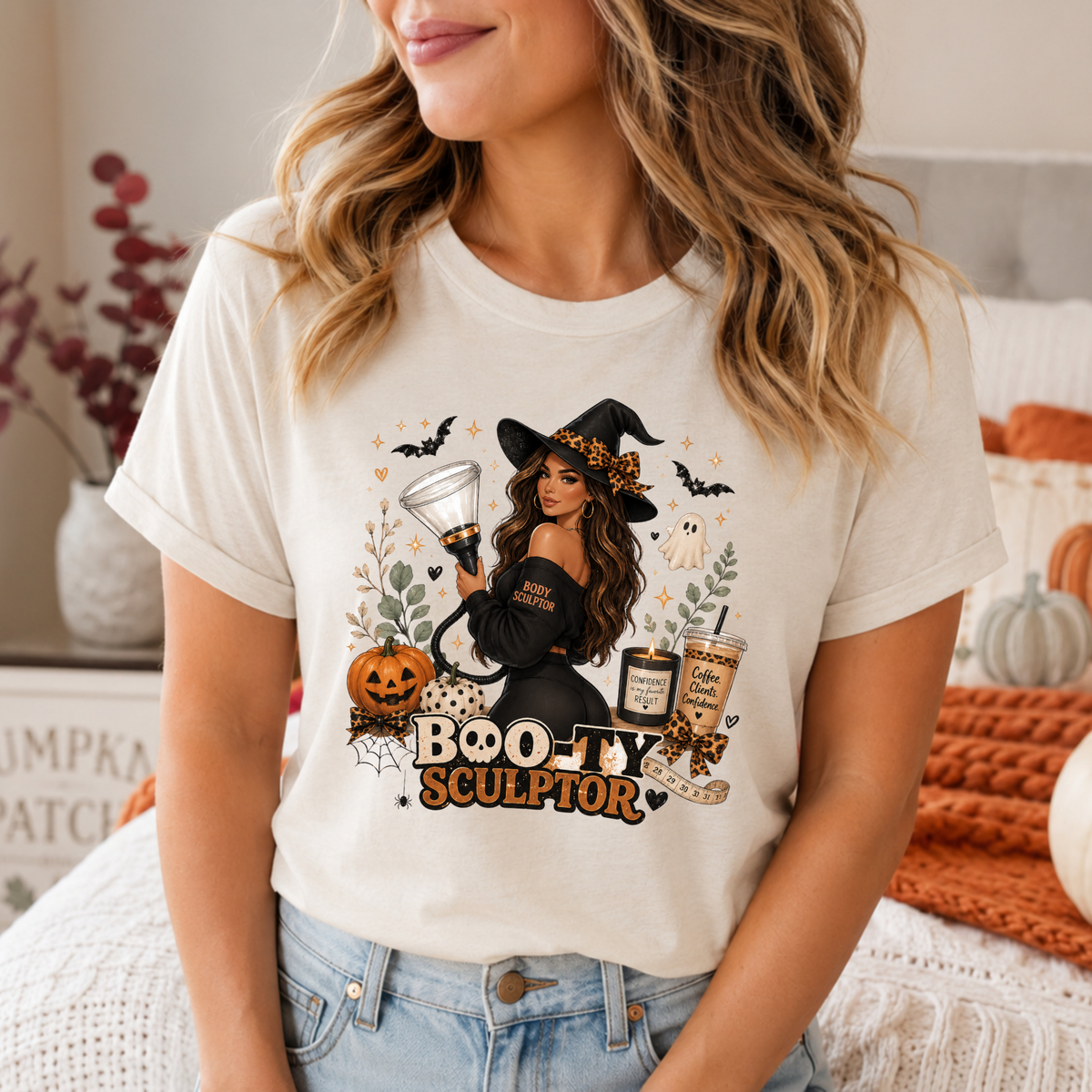 A woman wearing a cream-colored graphic t-shirt with a Halloween-themed design featuring a witch, pumpkins, bats, and the text 'BOO-TY SCULPTOR'.  Apparel and clothing for body contouring specialists and beauty professionals.