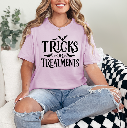 A woman wearing a light purple graphic t-shirt with 'TRICKS OR TREATMENTS' printed in black, paired with ripped light blue jeans, sitting on a white couch with a black and white checkered blanket. Clothing and apparel for beauty professionals.