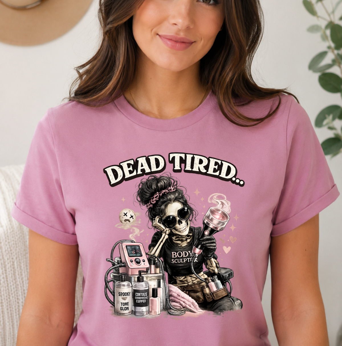 A woman wearing a pink graphic t-shirt with a cartoon skeleton holding vape devices and the text 'DEAD TIRED. Novelty shirts and apparel for body contouring specialists.