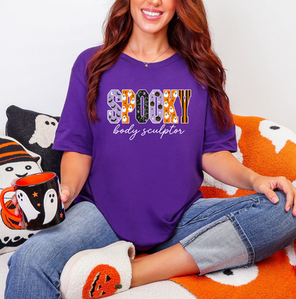 A woman wearing a purple graphic t-shirt with 'SPOOKY body sculptor' printed on the front, sitting on a cozy orange and white patterned couch and holding a black mug with a ghost design. Clothing and apparel for body contouring specialists and beauty professionals.