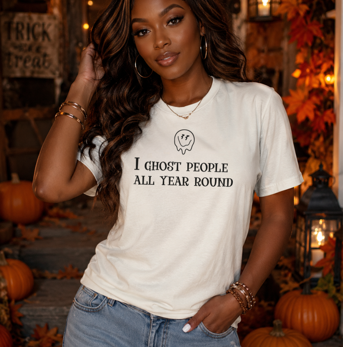 A woman wearing a white graphic t-shirt with the text 'I GHOST PEOPLE ALL YEAR ROUND' and a small ghost illustration, posed in a cozy, autumn-themed setting with pumpkins and fall decorations. Halloween graphic shirts and sweatshirts for women and beauty professionals.