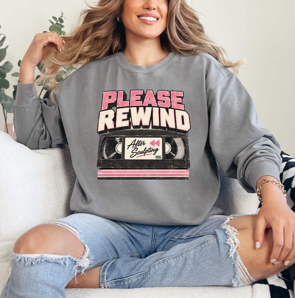 A woman sitting on a white couch wearing a gray crewneck sweatshirt with a retro graphic that reads 'PLEASE REWIND' above a VHS tape illustration. She is also wearing light-wash ripped jeans. Clothing, apparel and accessories for body contouring specialists and beauty professionals.