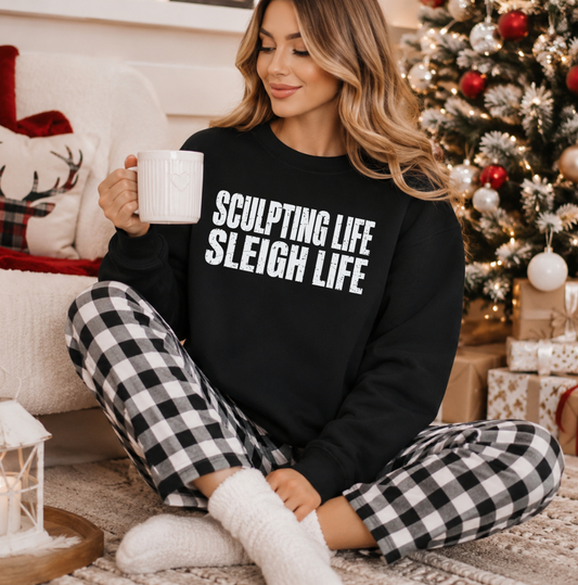 A woman sits on a rug in a cozy holiday setting, wearing a black sweatshirt with the white text 'SCULPTING LIFE SLEIGH LIFE' and black-and-white gingham pajama pants. She holds a white mug near a decorated Christmas tree and wrapped gifts. Holiday clothing and apparel for body contouring specialists and beauty professionals.