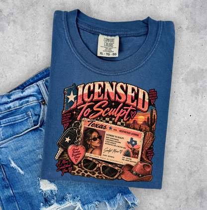 A folded blue graphic T-shirt laid flat on a light gray textured surface, styled with a pair of distressed blue jeans. The shirt features a large front graphic reading 'Licensed To Sculpt' with Texas-themed elements including a license ID card, cowboy boots, sunglasses, keys, and a heart-shaped tag. Clothing, apparel and accessories for body contouring specialists and beauty professionals.