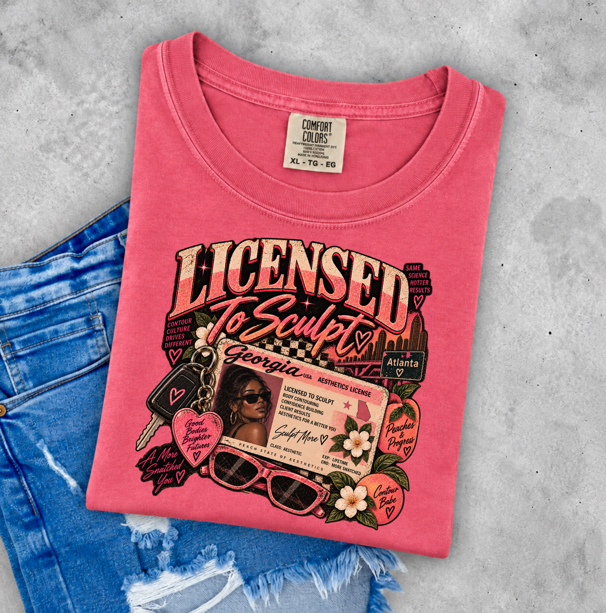 A folded pink graphic T-shirt is laid flat on a light gray textured surface, partially covered by blue denim jeans. The shirt features a large retro-style graphic with the words 'Licensed to Sculpt,' a Georgia license-plate design, sunglasses, flowers, and small decorative elements. Clothing, apparel and gifts for body sculptors, estheticians, and beauty professionals.