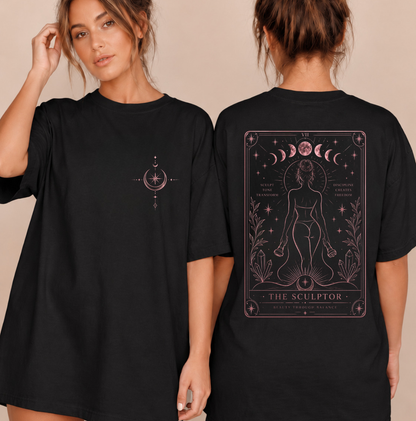 Two models wearing a black oversized T-shirt dress, showing the front and back designs. The front features a small pink celestial graphic on the left chest, and the back features a large pink tarot-style card graphic titled 'THE SCULPTOR' with moon phases, a nude female figure, and botanical details. Clothing, apparel and accessories for body contouring specialists, and beauty professionals.