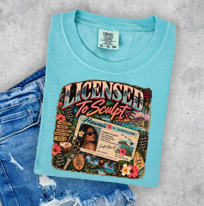 A light aqua blue graphic T-shirt laid flat on a gray concrete surface, styled with distressed blue jeans. The shirt features a large colorful front graphic with the words 'Licensed To Sculpt' and a Hawaii-themed design. Clothing, apparel and accessories for body contouring specialists and beauty professionals.