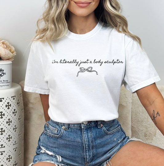 A woman wearing a white short-sleeve graphic T-shirt with the text 'i'm literally just a body sculptor' and a simple line drawing of a body contouring, paired with denim shorts in a cozy indoor setting. Clothing and apparel for body contouring specialists and beauty professionals.
