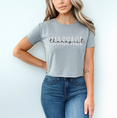 A woman wearing a heather gray short-sleeve t-shirt with the text 'therapist' printed across the chest in black script and a faded white block lettering version of the same word. She is wearing blue jeans and posing against a light blue wall. Clothing and apparel for massage therapists and beauty professionals.