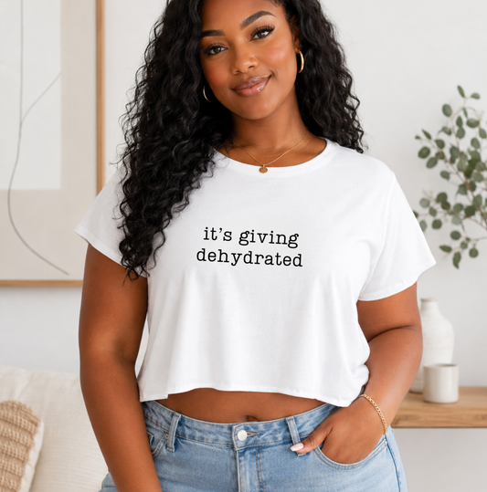 A woman wearing a white cropped graphic t-shirt with the text 'it's giving dehydrated' in black lowercase letters, paired with light blue jeans in a bright, modern indoor setting. Shirts and apparel for body contouring specialists, massage therapists and beauty professionals.