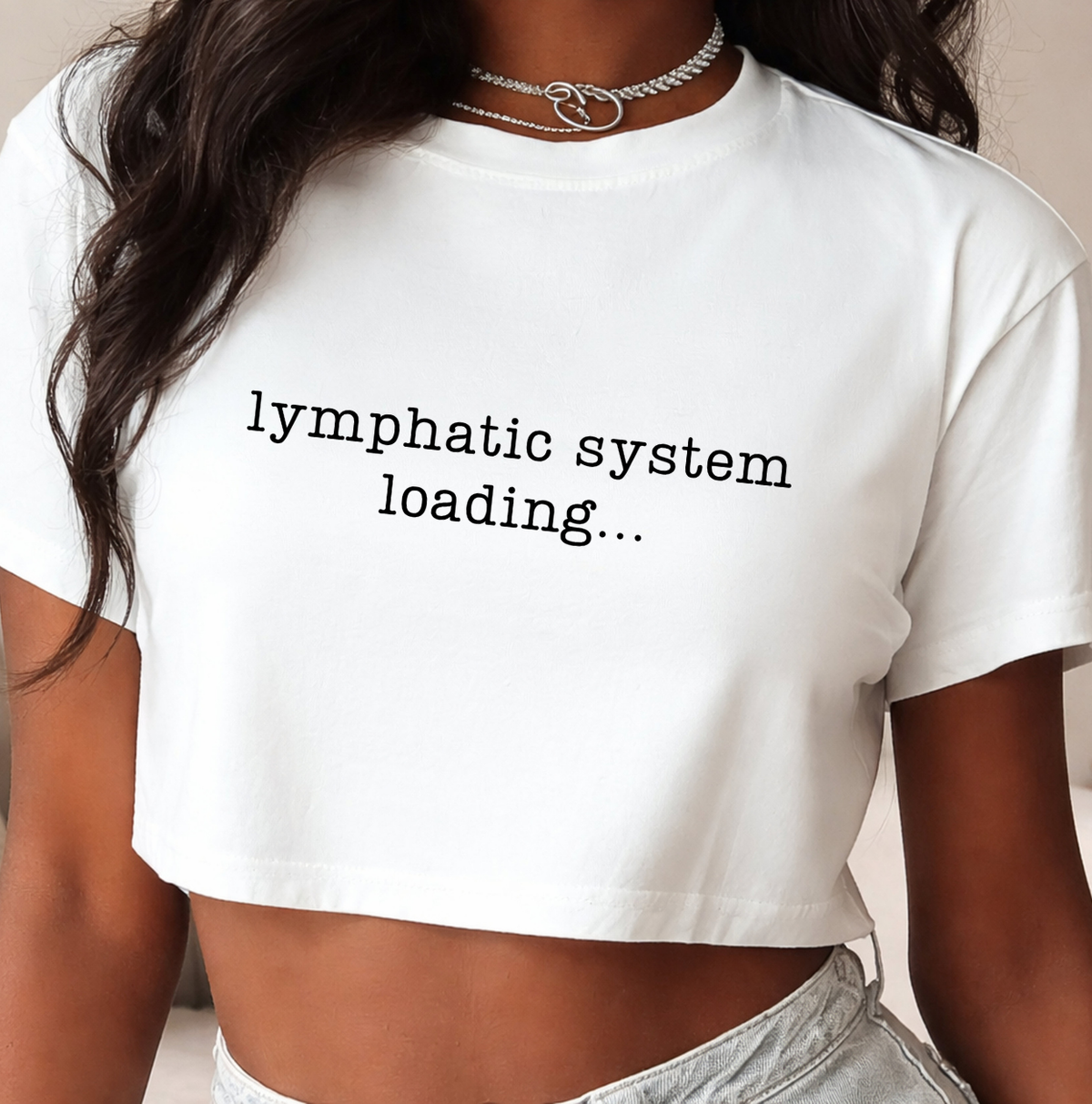 A close-up of a person wearing a white cropped T-shirt with the text 'lymphatic system loading. Novelty shirts and clothing for body contouring specialists, massage therapists, nurses, and beauty professionals.