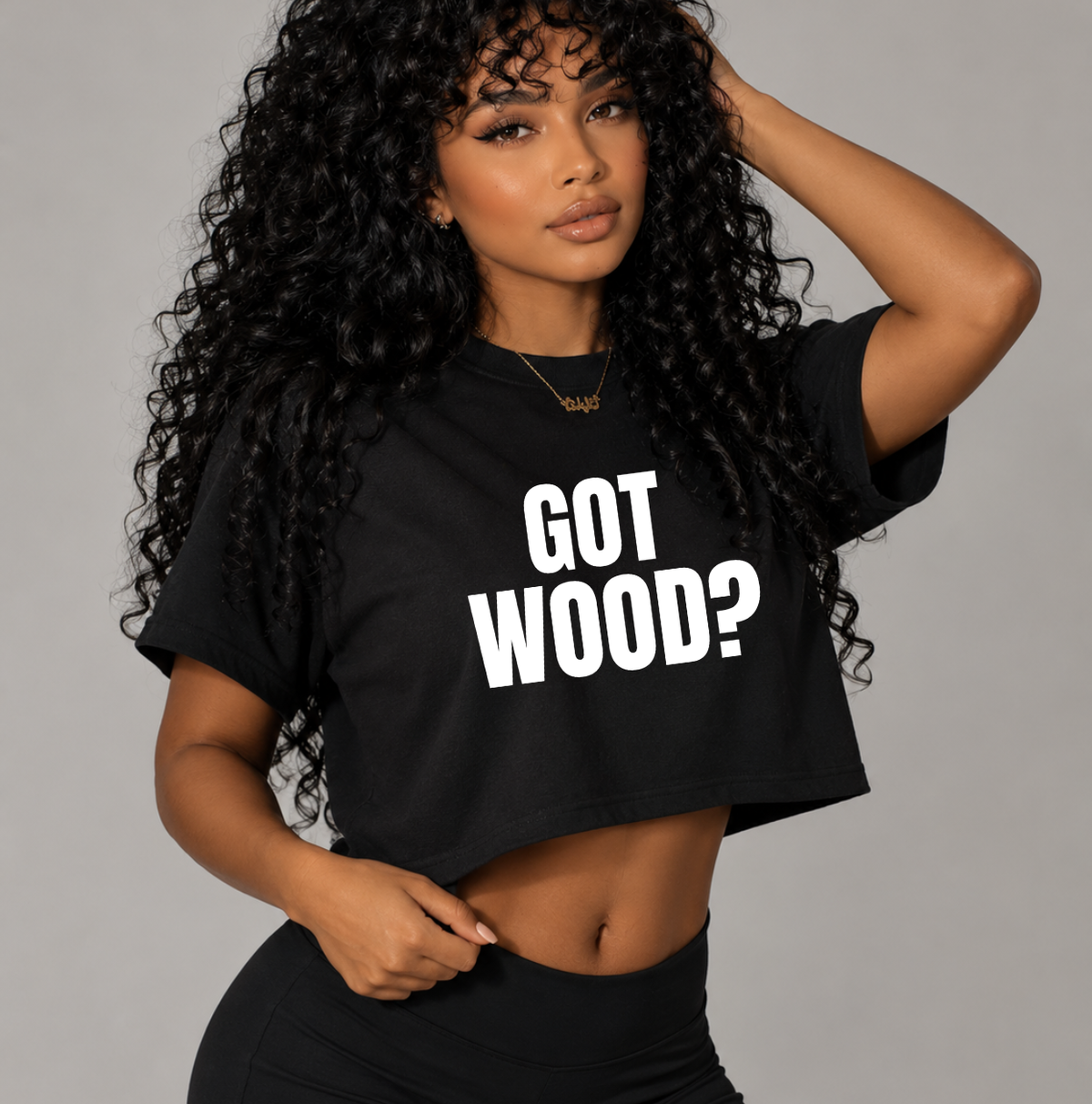 A woman with long, curly black hair models a black crop top t-shirt featuring the bold white text 'GOT WOOD?'. She is posing against a solid gray studio background. Graphic t-shirts and apparel for body contouring specialists, maderotherapy and wood therapy specialists, and beauty professionals.