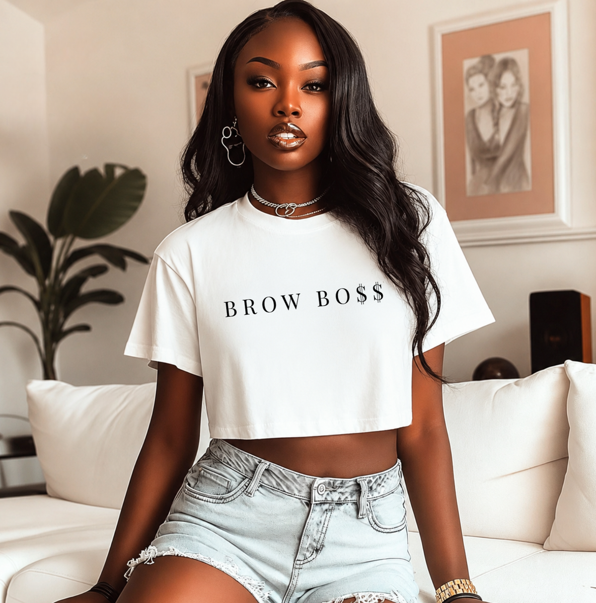 A woman wearing a white cropped t-shirt with 'BROW BOSS' printed in black across the chest, paired with light-wash denim shorts, sitting on a white couch in a modern living room setting. Gifts & apparel for PMU artists.