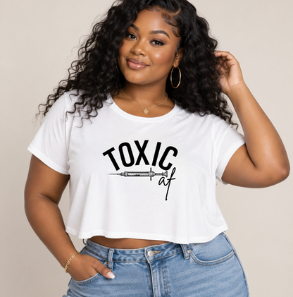 A woman wearing a white cropped t-shirt with a black graphic that reads 'TOXIC af' with a syringe illustration, paired with blue jeans, against a solid beige background. Clothing, apparel and accessories for nurse injectors and beauty professionals.