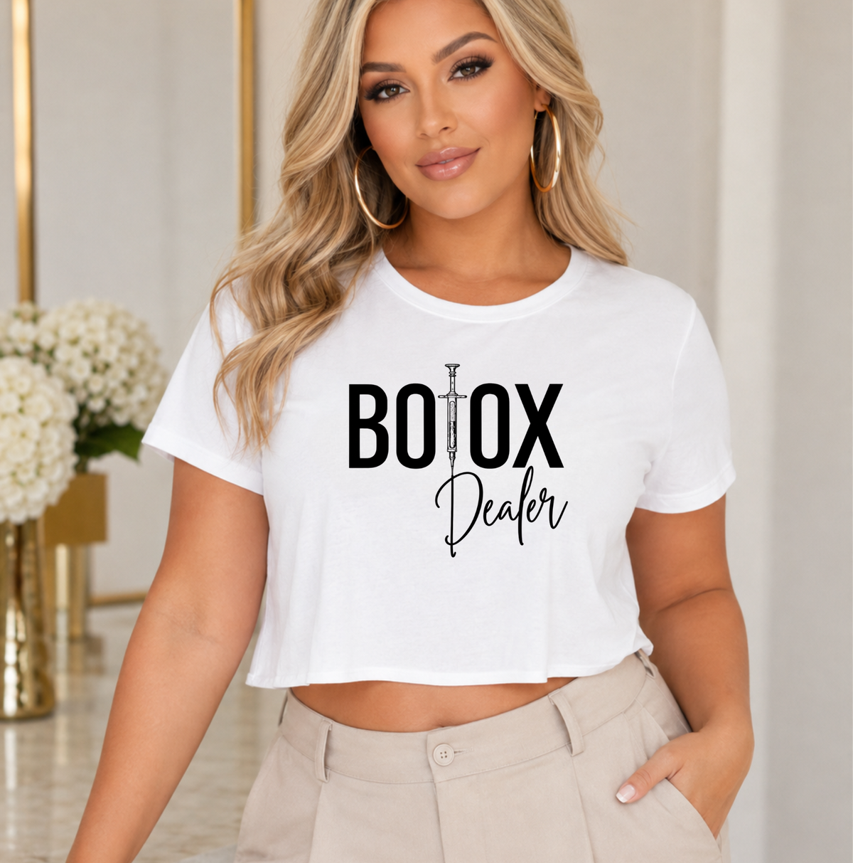 A woman wearing a white cropped T-shirt with a black graphic that reads 'BOX Dealer' and light beige pants, standing in an indoor setting with a blurred background. Clothing, apparel and accessories for nurse injectors and beauty professionals.