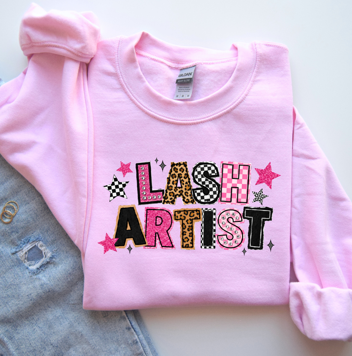 A folded light pink long-sleeve sweatshirt with a colorful 'LASH ARTIST' graphic on the front, styled on a white surface next to a light blue denim skirt. Gifts and apparel for lash artists.