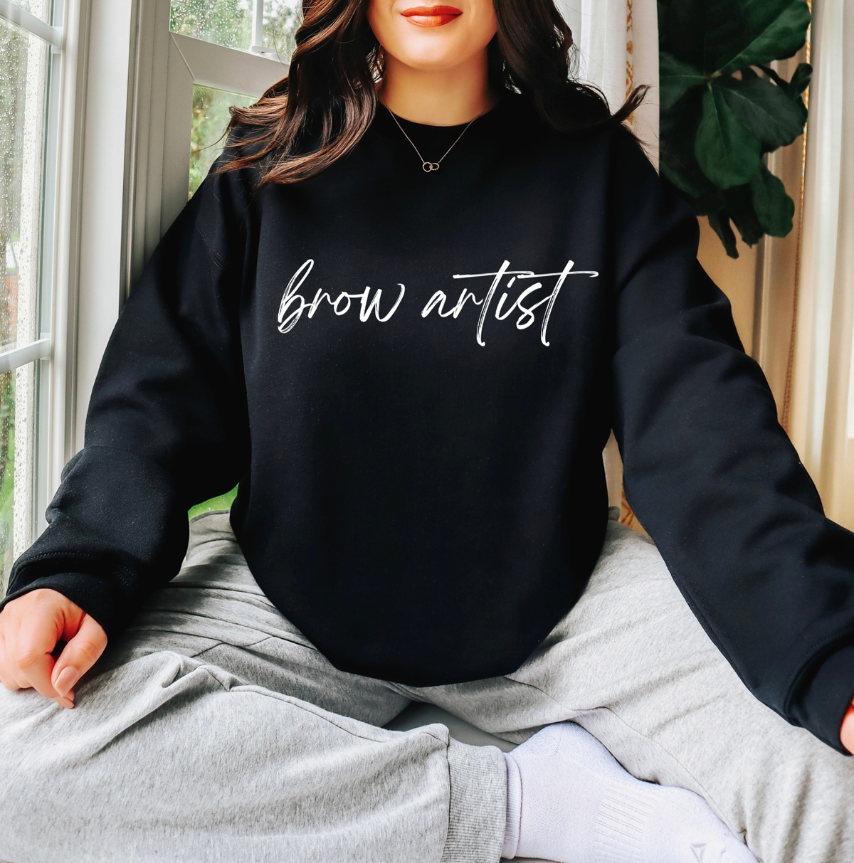 A woman wearing a black crewneck sweatshirt with 'brow artist' printed in white script across the chest, paired with light gray sweatpants and white socks, sitting in front of a window. Gifts and apparel for PMU and brow artists.
