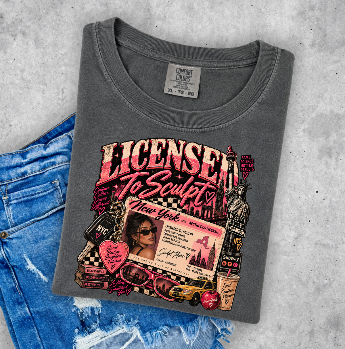 A folded dark gray graphic t-shirt with a colorful 'Licensor to Sculpt' New York graphic print, styled on a light gray concrete background next to a pair of distressed blue jeans. Clothing and gifts for beauty professionals.