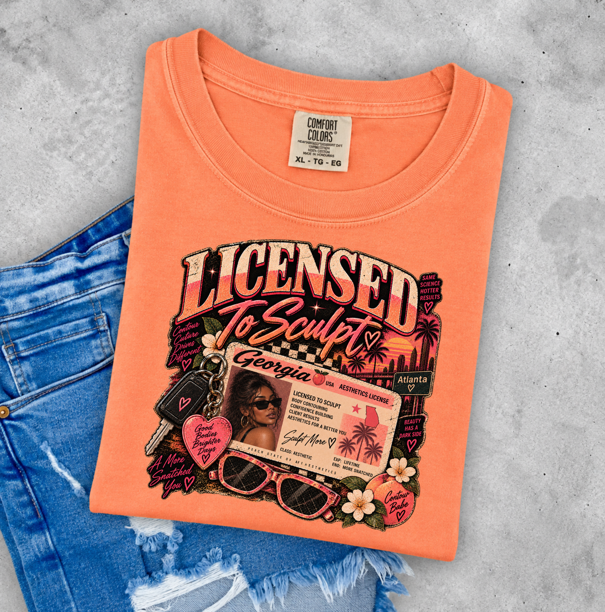 An orange crewneck t-shirt with a colorful graphic print reading 'LICENSED to Sculpt' and featuring a cartoon character, sunglasses, and keys, styled flat-lay on top of blue jeans against a gray concrete background. Clothing and gifts for beauty professionals.