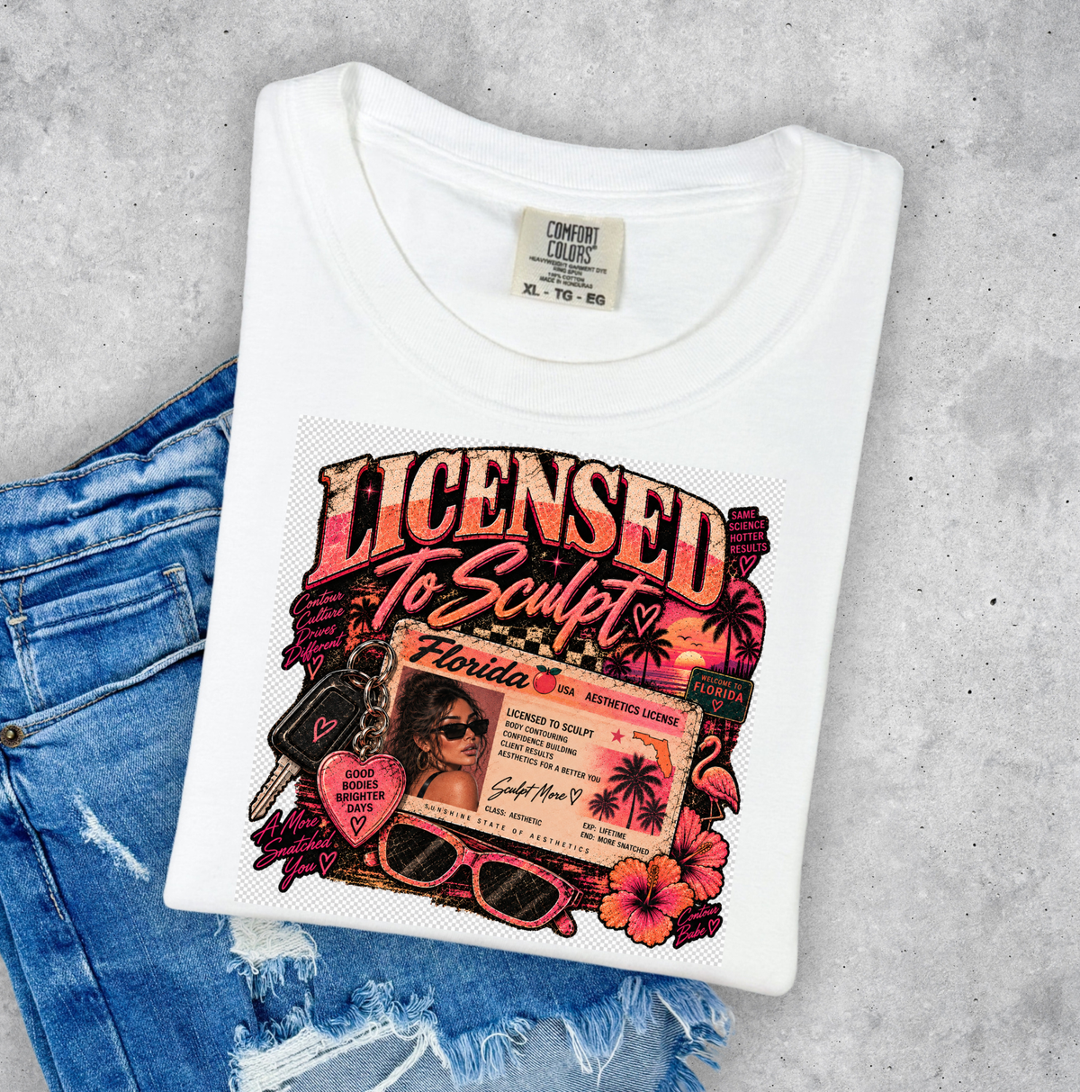 A folded white graphic T-shirt displayed on top of distressed blue jeans against a light gray concrete-style background. The shirt features a colorful retro-style print reading 'Licensed to Sculpt' with floral, sunglasses, keys, and a Florida license-idea collage. Clothing for body contouring specialists.