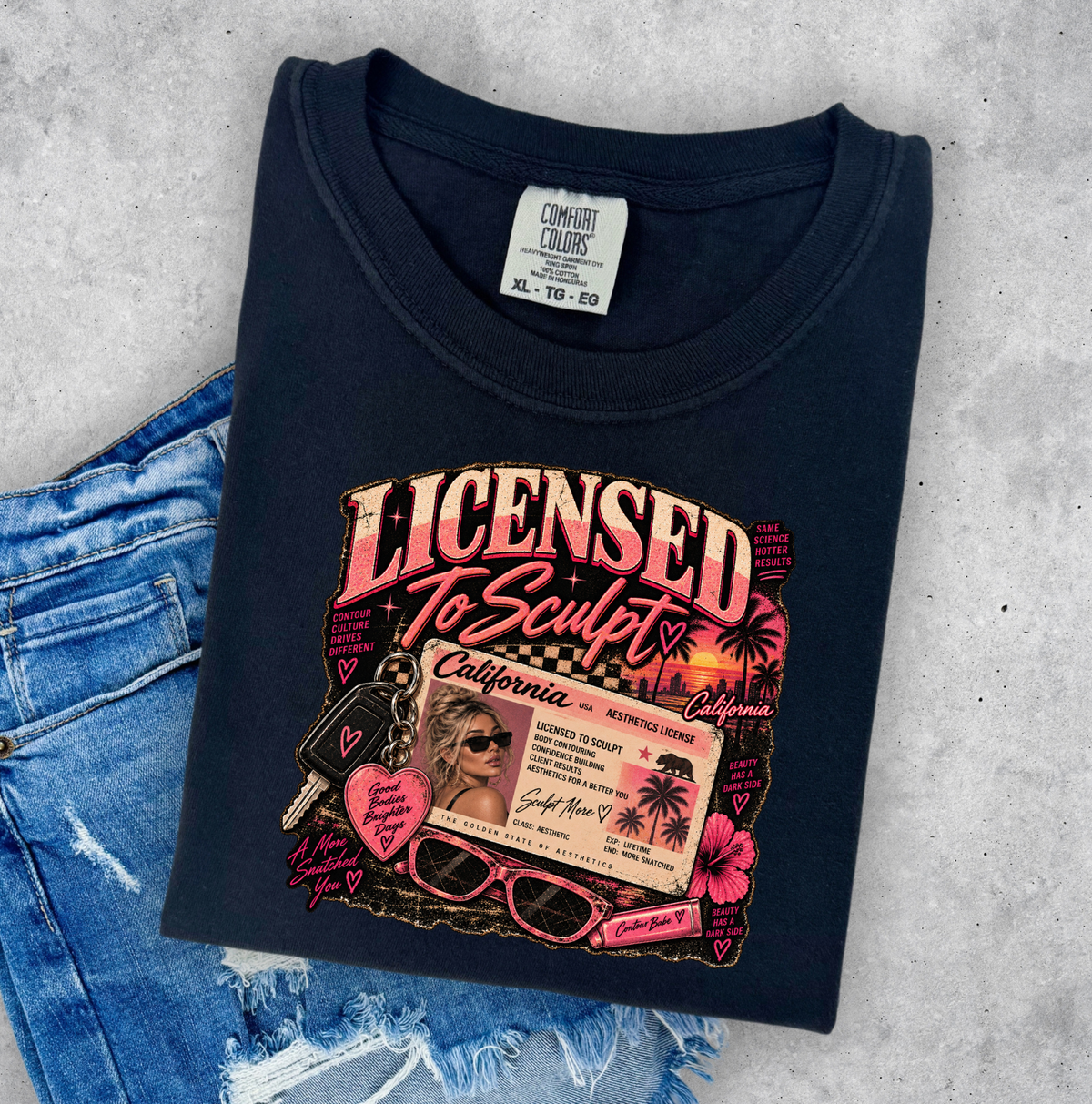 A folded black graphic t-shirt with a pink and gold 'Licensed To Sculpt' print, styled flat-lay on a gray concrete background next to a pair of distressed blue jeans. Clothing for beauty professionals.