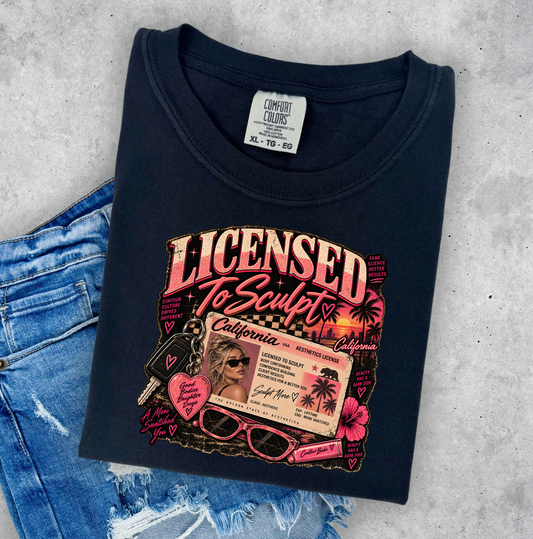 A folded black graphic t-shirt with a pink and gold 'Licensed To Sculpt' print, styled flat-lay on a gray concrete background next to a pair of distressed blue jeans. Clothing for beauty professionals.