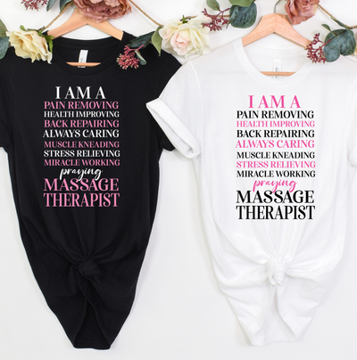 Two t-shirt dresses displayed side by side on wooden hangers against a white background, decorated with artificial flowers at the top. The left dress is black with pink text, and the right dress is white with pink and black text. Clothing, apparel, and accessories for massage therapists and beauty professionals.