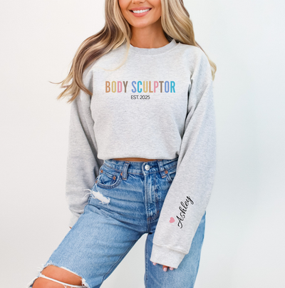 A woman wearing a light gray cropped sweatshirt with 'BODY SCULPTOR EST 2025' printed across the chest in rainbow colors, paired with distressed blue jeans. The sweatshirt also has 'Ashley' and a small pink heart printed on the left sleeve.