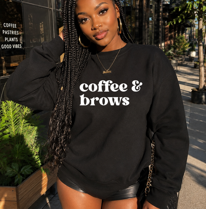 A woman wearing a black crewneck sweatshirt with 'coffee & brows' printed in white across the chest, photographed outdoors on a city sidewalk near a coffee shop. Gifts & apparel for PMU artists.